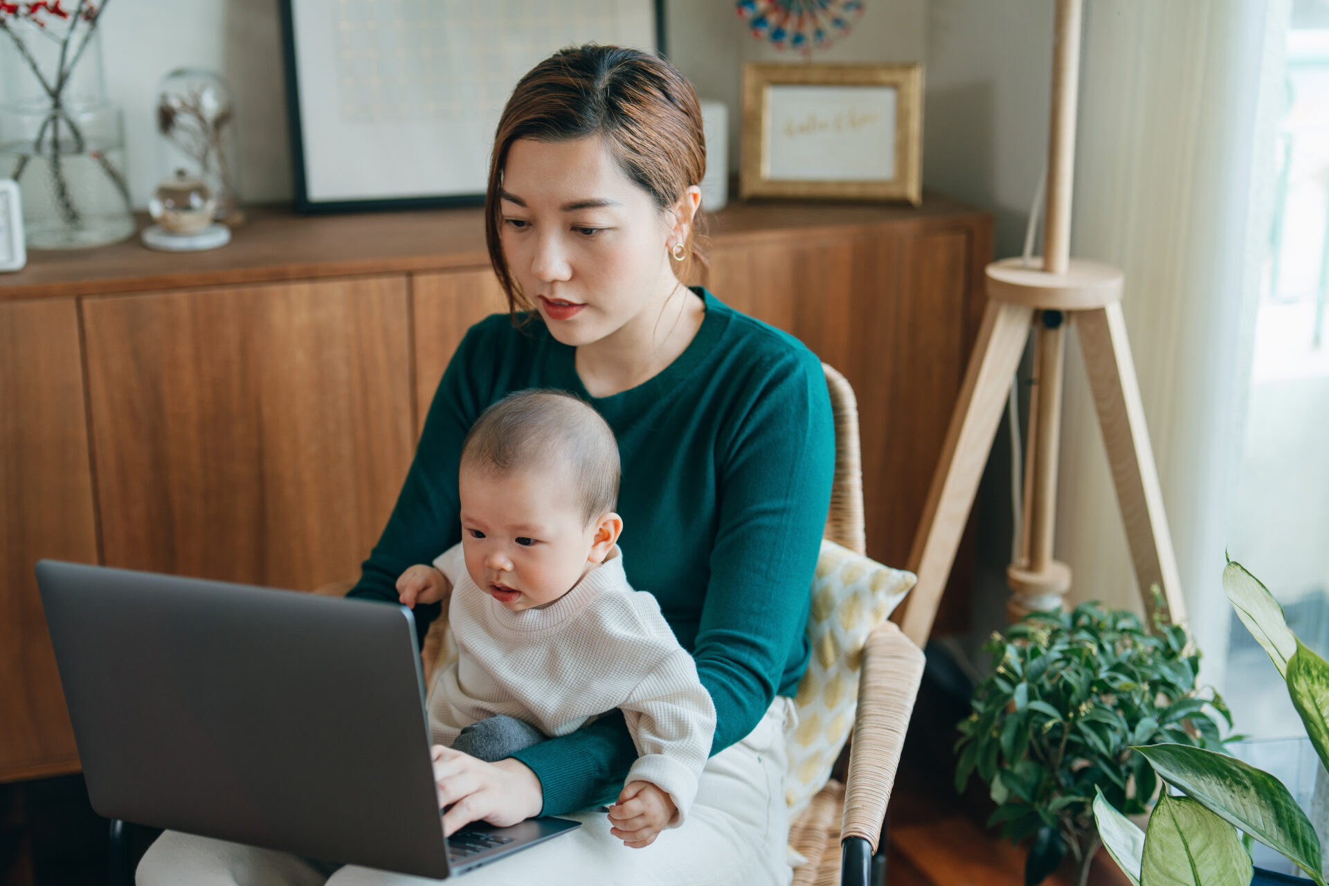 Young Asian mother managing home finances, banking and finance with laptop while taking care of her baby girl sitting on her lap. Multi-tasking working mother managing home issues and childcare at home